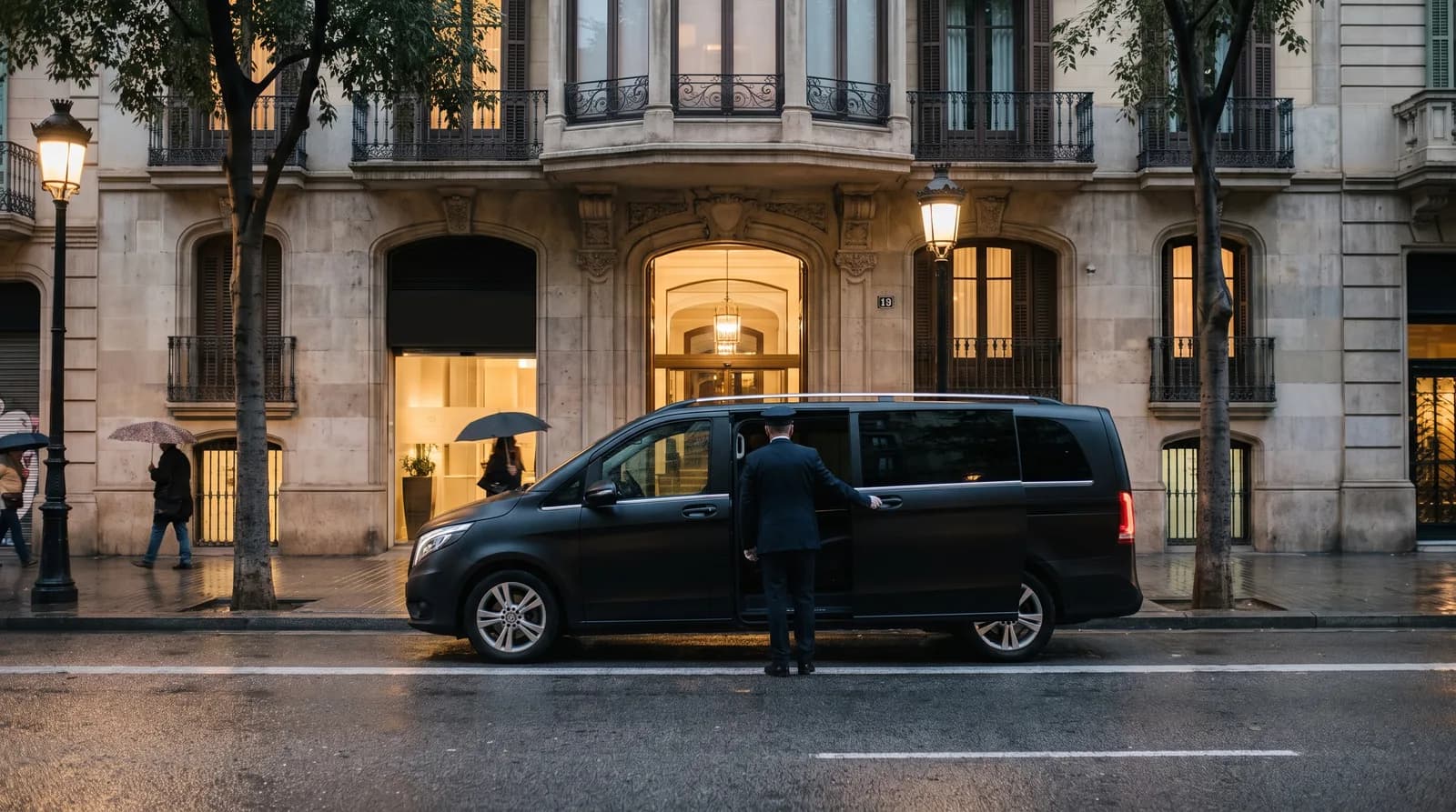 Chófer abriendo la puerta lateral de un monovolumen negro ante un edificio del Eixample de Barcelona al anochecer